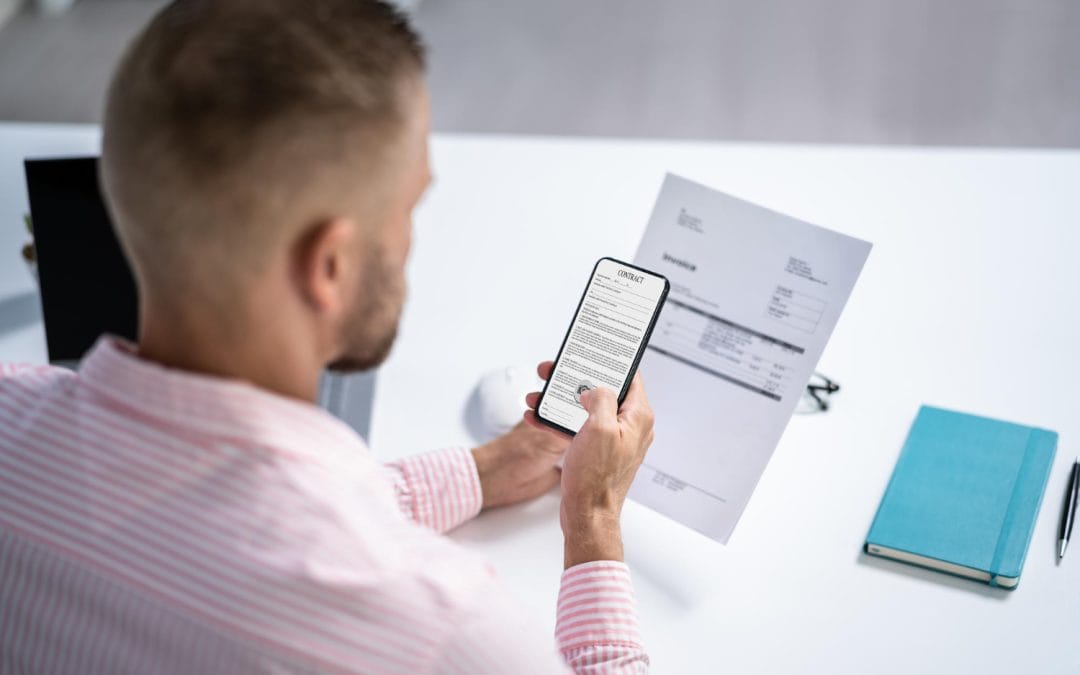 Man at desk holding a smartphone and an invoice, with a blue notebook and laptop nearby.