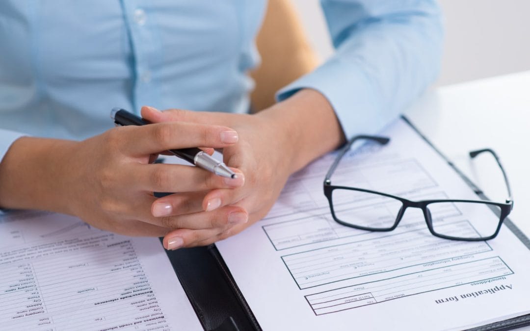 Close-up of hands holding a pen over application forms and a pair of eyeglasses on a desk.