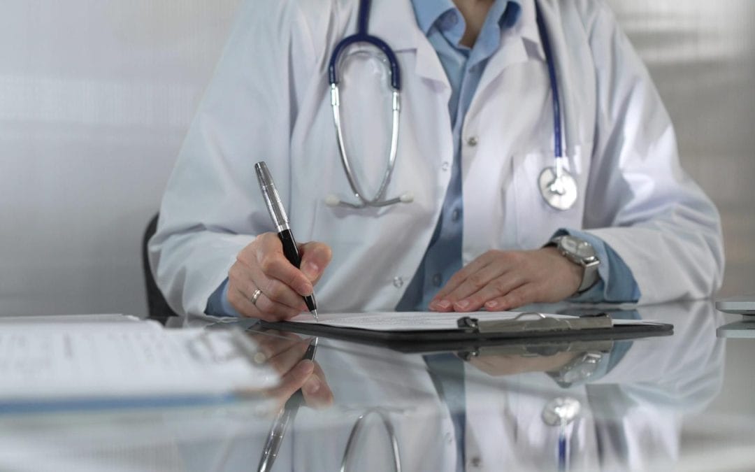 Doctor in white coat writing on a clipboard at a facis level 3 desk with a stethoscope around their neck.